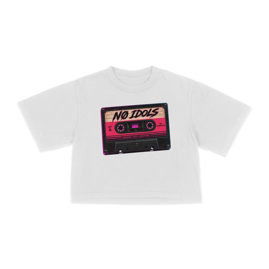 A white short-sleeve crop top t-shirt featuring a graphic print of a retro cassette tape in pink, black, and beige with the text 'NO IDOLS' written on it, displayed against a solid white background.